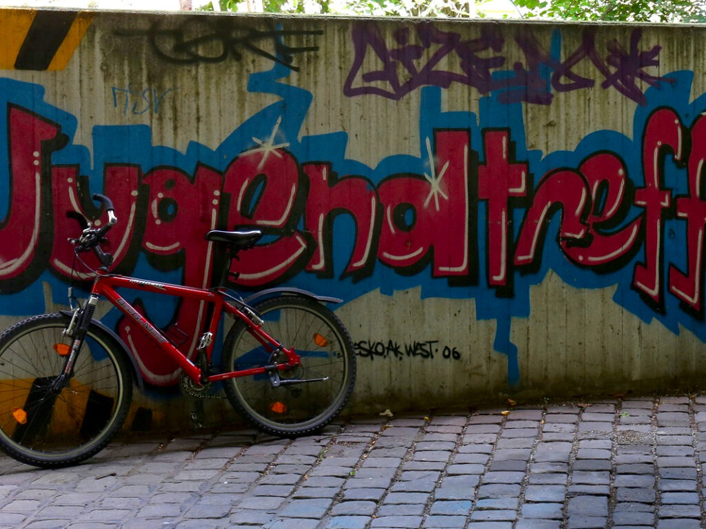 Eine Holzwand mit dem roten Graffiti-Schriftzug "Jugendtreff" an der links vorn ein rotes Fahrrad lehnt