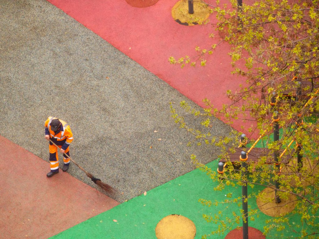 Luftaufnahme eines Mannes in einer orange-schwarzen Uniform der einen bunten Hartplatz kehrt.
