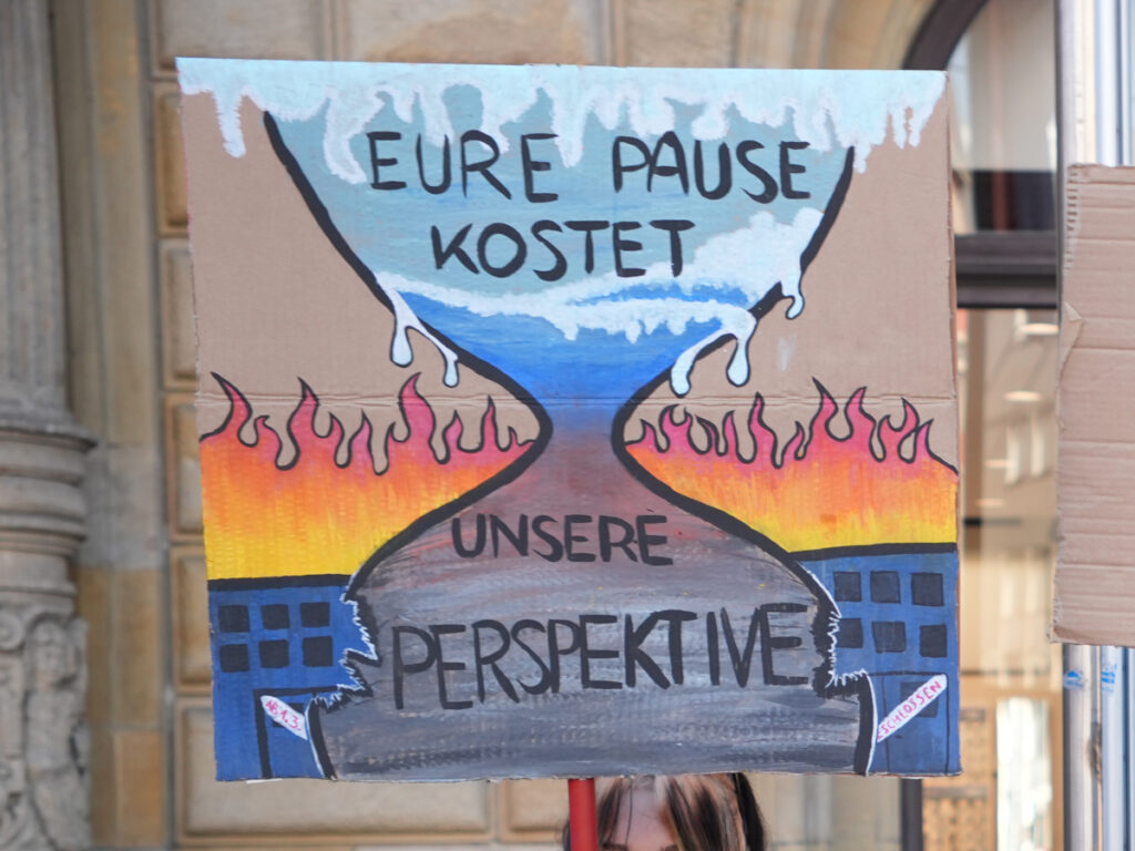 Handgemaltes Protestplakat mit der Aufschrift: „Eure Pause kostet unsere Perspektive“. Es zeigt eine mit schmelzendem Eisland gefüllte Sanduhr über einer brennenden Stadt.