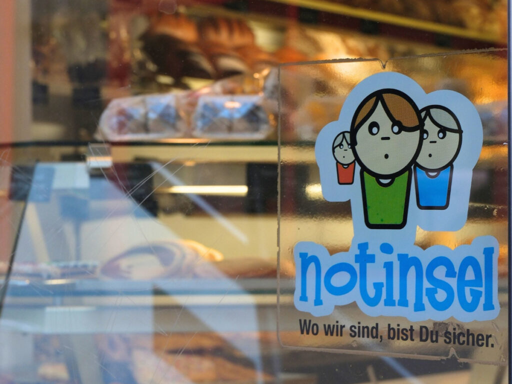 Im Hintergrund die warm beleuchte Auslage einer Bäckereifilliale, im Vordergrund rechts der "Notinsel"-Aufkleber auf der Scheibe. Der Aufkleber zeigt drei comichaft stilisierte Kinder, den großen Schriftzug "Notinsel" und die kleinere Unterzeile "Wo wir sind, bist Du sicher."